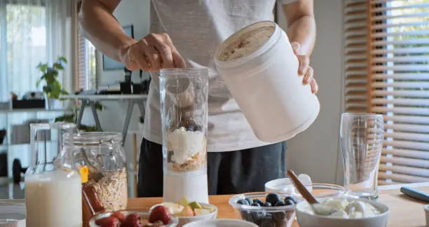 A man making a protein shake in his kitchen in a white shirt 