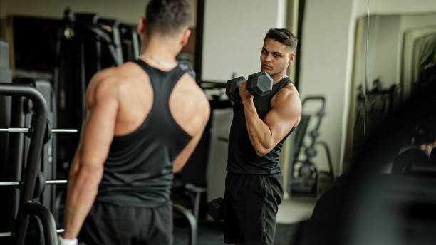 A man working out in the gym with a mirror looking at himself doing a bicep curl 