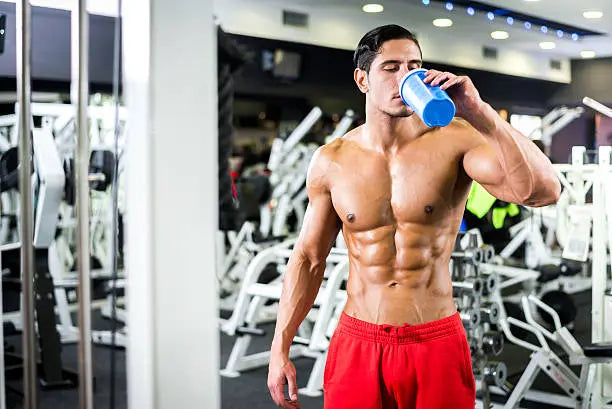 Man drinking pre workout in a gym in red shorts. He is very fit and has large muscles due to working out and training. 