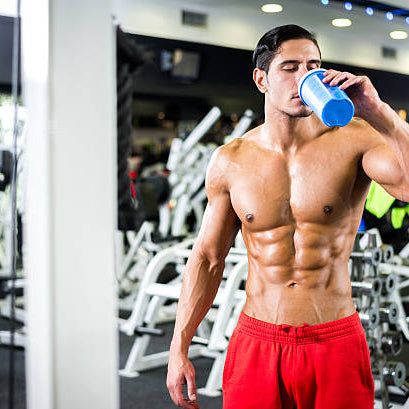 Man drinking pre workout in a gym in red shorts. He is very fit and has large muscles due to working out and training. 