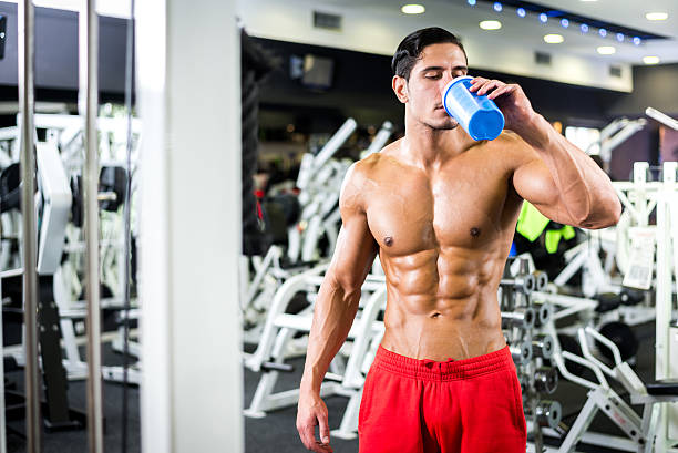 Man drinking pre workout in a gym in red shorts. He is very fit and has large muscles due to working out and training. 